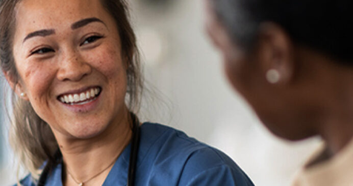 A smiling healthcare professional in blue scrubs, wearing a stethoscope, engaging in a conversation with a patient.